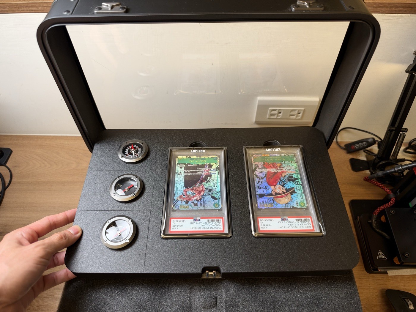 The Kalijoy K CASE front panel folded down, showing three round dials on the left and two One Piece PSA slabs in Kalijoy holders.