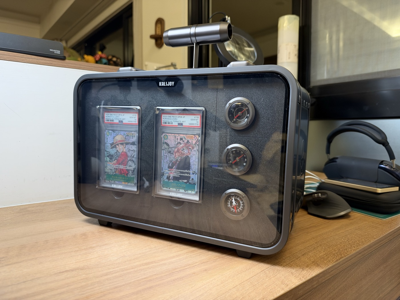 The Kalijoy K CASE closed on a desk, its front panel displaying two One Piece PSA slabs beside three round dials.